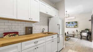 A modern kitchen with white cabinets, a wooden countertop, and a built-in sink. There is a refrigerator, a dining area with a table and chairs, and a painting on the wall.