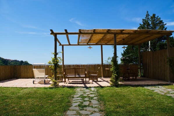 A wooden pergola with a bench and table, surrounded by a wooden fence and a stone pathway. - AntiquaeLunae - Callistemon (Fosdinovo)