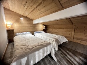 Una habitación con dos camas, paredes de madera, techo inclinado y una mesita de noche pequeña con una lámpara.