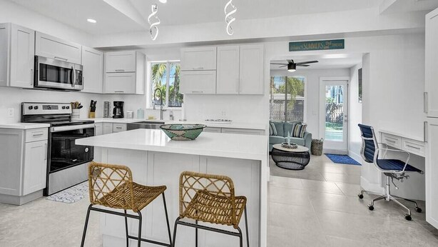 A modern kitchen with white cabinetry, a central island, two wicker bar stools, a dining area with a blue sofa, and a ceiling fan. - Luxury & Comfy Pool Getaway ~ Mins to Beaches & DT (Fort Lauderdale)