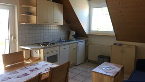 Une cuisine compacte avec des meubles blancs, un crédence carrelée, une cuisinière et un évier. Une fenêtre laisse entrer la lumière naturelle, et il y a une petite table avec des chaises.