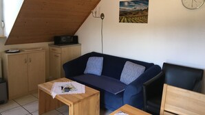 Une pièce chaleureuse avec un plafond en pente, un canapé bleu foncé agrémenté de coussins, une table en bois, un petit meuble et une horloge au mur.