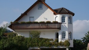 Une maison à deux étages avec un balcon, des murs blancs et un toit brun.