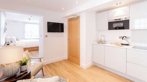 A modern, well-lit room with a bed, a TV, a wooden door, a kitchen with white cabinets, and a small table with a lamp and a plant. - Snug Cuppin Street Apartment (Chester)