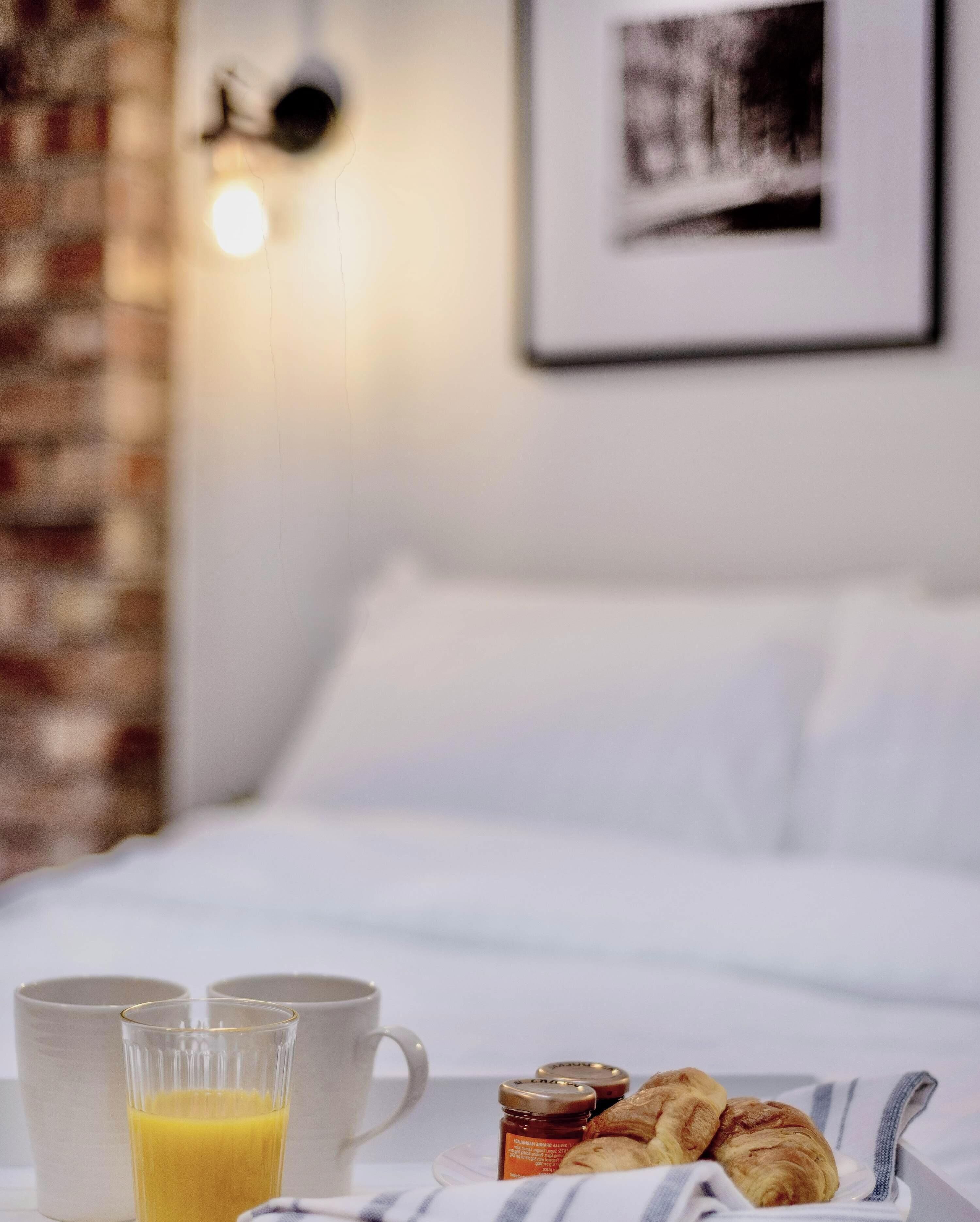 Una bandeja de desayuno bien presentada con cruasanes, un vaso de zumo de naranja y una taza, sobre una cama con sábanas blancas y un cuadro en la pared.