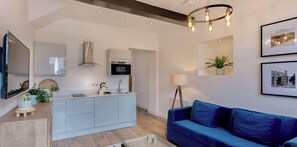 A modern living room with a blue sofa, a kitchen area with a built-in oven, a large mirror, and a ceiling light fixture. - Grosvenor Street Stylish 1 Bed Flat (Chester)
