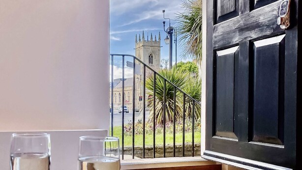 Un balcon avec deux verres de vin blanc, une vue sur une église et une porte noire sur laquelle est fixé un téléphone.