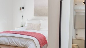 A bedroom with a bed, a bedside table, a bench, a wall-mounted lamp, and a painting.