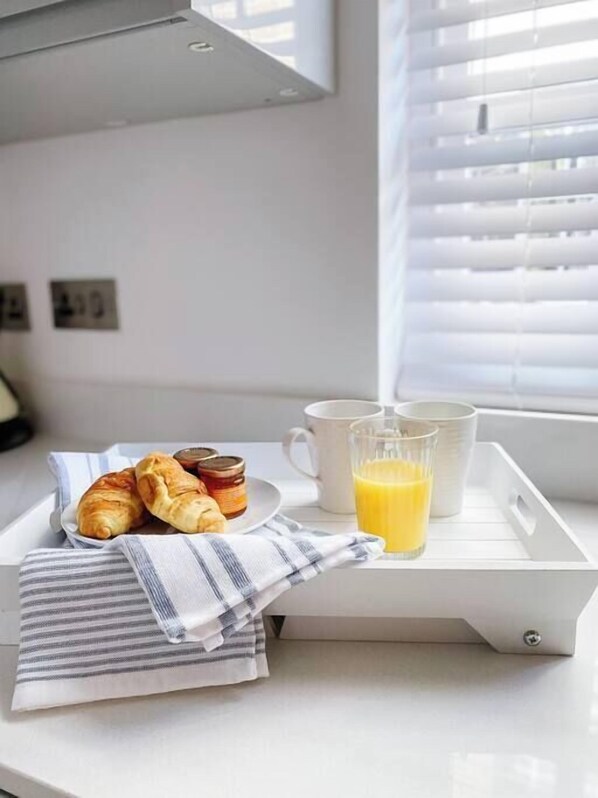A tray with croissants, a glass of orange juice, and two white cups on a white countertop near a window with blinds.