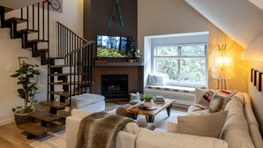 Una sala de estar moderna con chimenea, una escalera y un amplio ventanal con vistas a árboles.