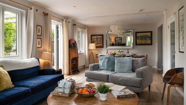 Ein gemütliches Wohnzimmer mit einem blauen Sofa, einem grauen Sofa und einem Couchtisch aus Holz. An den Wänden befinden sich gerahmte Bilder, eine Lampe und ein Fenster mit Vorhängen.