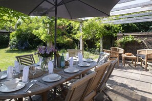Un área de comedor al aire libre con una mesa preparada para seis personas, rodeada de sillas de madera y una sombrilla.