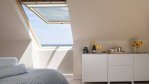 Ein Schlafzimmer mit Oberlicht, einem Bett mit grauer Decke, einer weißen Kommode und einem kleinen Tisch mit Flasche, Gläsern und einer Vase mit Sonnenblumen.