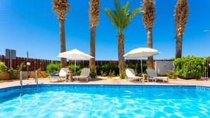 A swimming pool with lounge chairs and umbrellas, surrounded by palm trees and a wooden fence. - Queens Paradise 1 - Three Bedroom Villa, Sleeps 6 (Peyia)