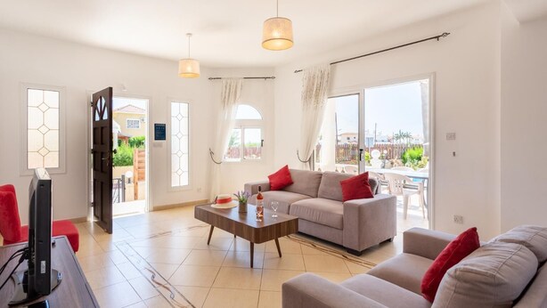 A spacious living room with a sofa, a coffee table, and a television. There are large windows and a balcony visible. - Queens Paradise 1 - Three Bedroom Villa, Sleeps 6 (Peyia)