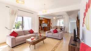 A spacious living area with two sofas, a coffee table, and a television. A kitchen is visible in the background. - Queens Paradise 1 - Three Bedroom Villa, Sleeps 6 (Peyia)