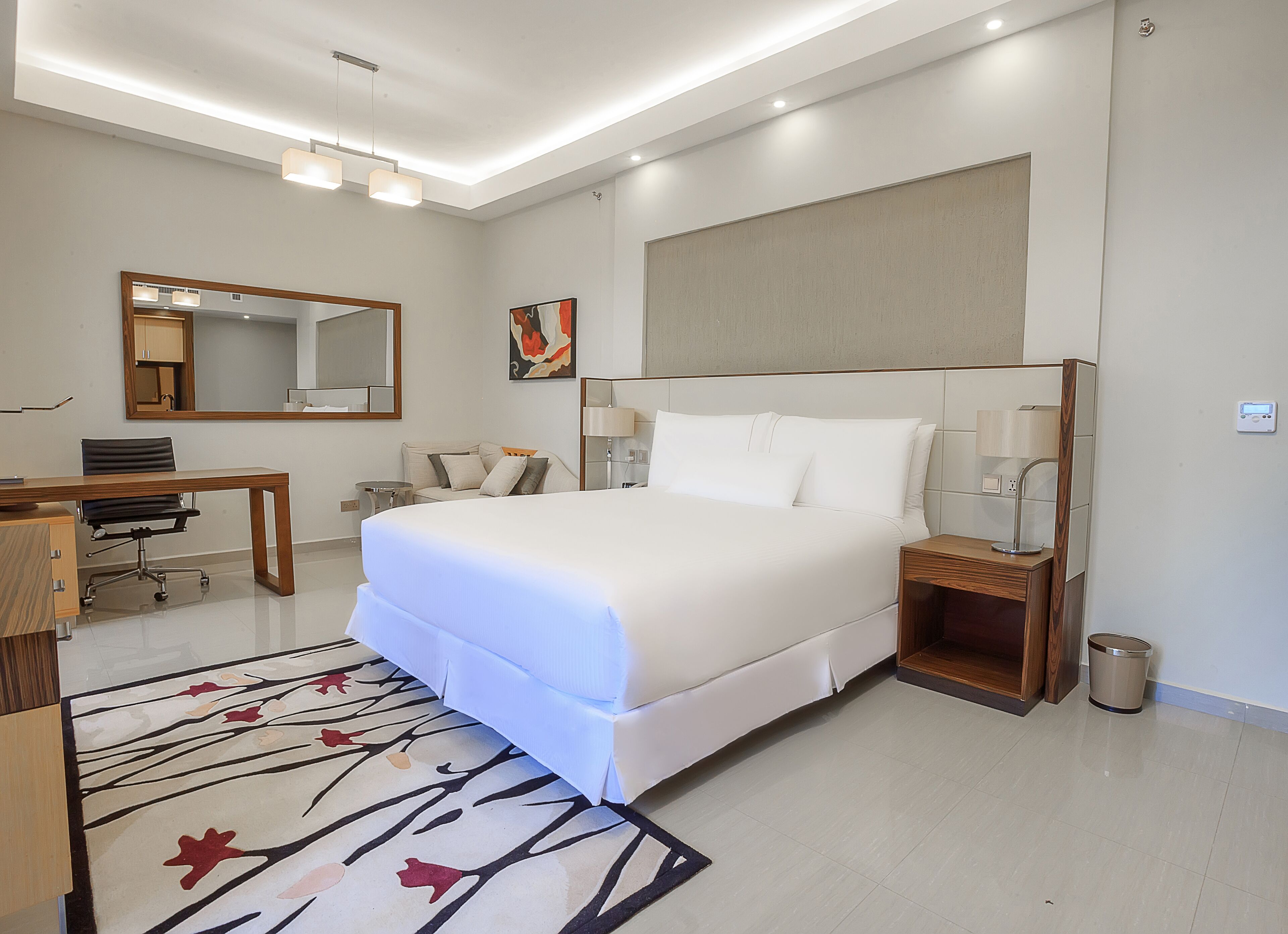 A modern hotel room with a large bed, a desk, a chair, a mirror, and a bedside table with a lamp.