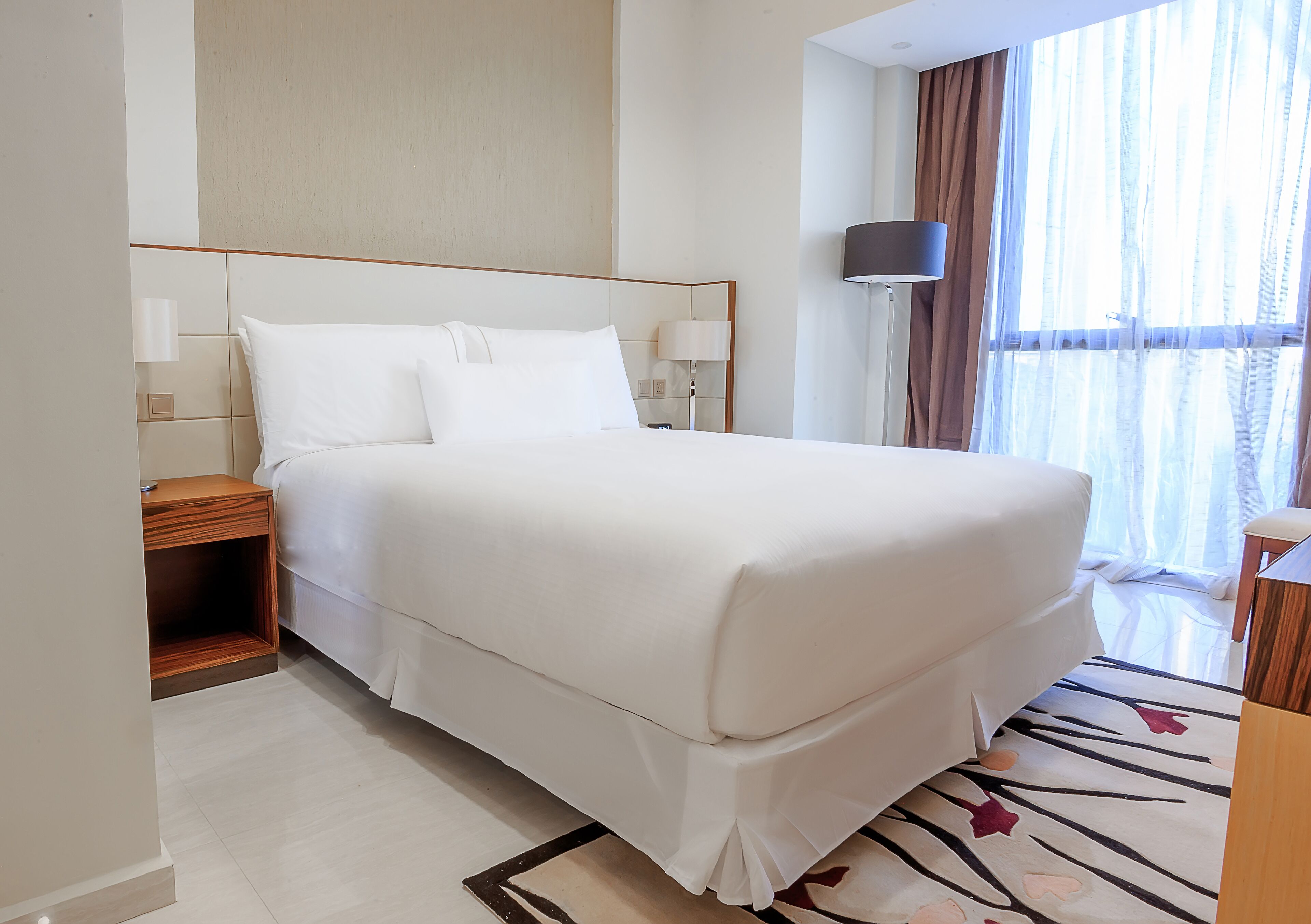 Una cama bien hecha con sábanas blancas, una mesita de noche con lámpara, y un amplio ventanal con cortinas de gasa.