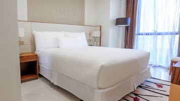 Una cama bien hecha con sábanas blancas, una mesita de noche con lámpara, y un amplio ventanal con cortinas de gasa.