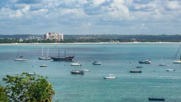 A coastal view with multiple boats anchored in calm waters, a distant beach, and a cluster of buildings in the background.