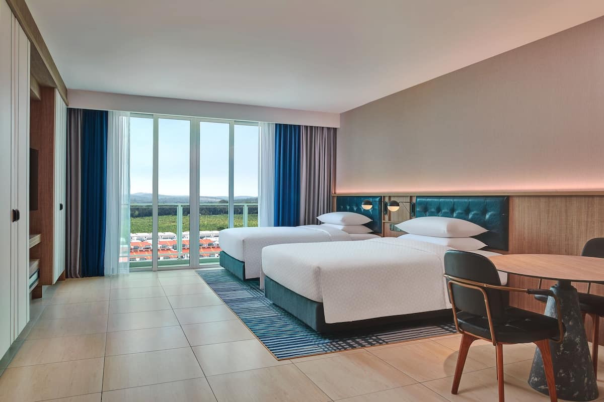 A hotel room with two beds, a dining table, and a view of a cityscape.