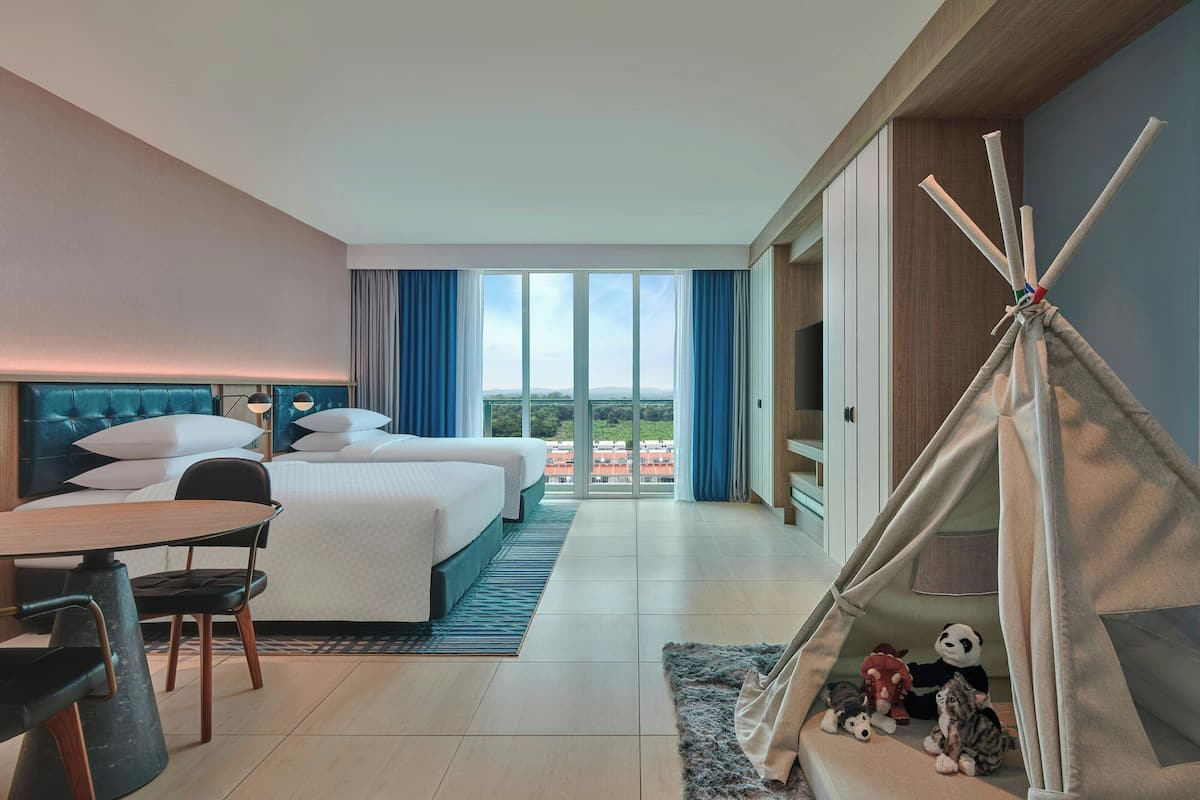 A hotel room with a bed, a desk, a chair, a dining table, a view of the city, and a teepee with stuffed animals.