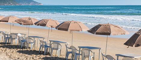 Una playa con filas de sillas y mesas de plástico bajo sombrillas marrones, frente al océano.