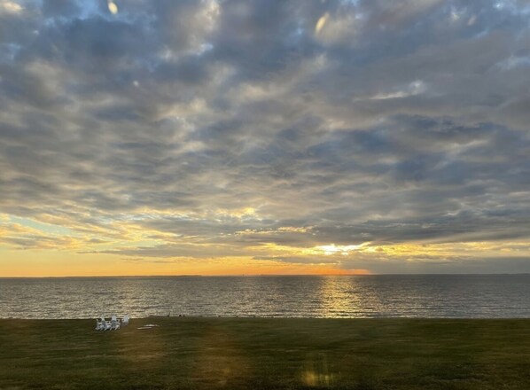 Una playa con un atardecer, césped y algunas sillas blancas.