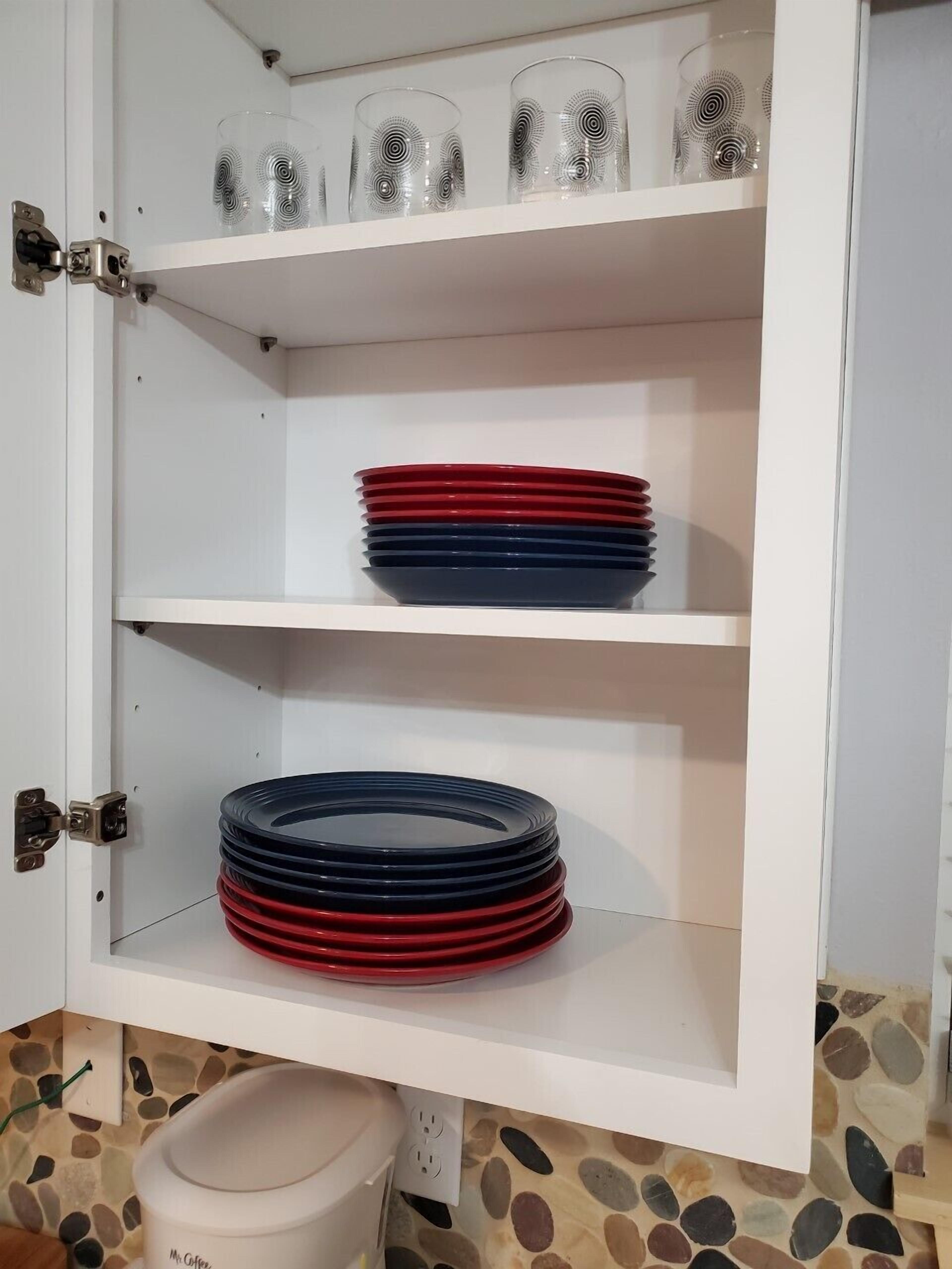 Ett vitt skåp med tre hyllor. På den översta hyllan står genomskinliga glas med ett spiralmönster, på den mellersta hyllan ligger staplar av röda och blå tallrikar, och på den nedersta hyllan ligger en hög med svarta tallrikar.