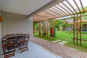 A covered patio area with a table and chairs, a brick pathway, and a grassy yard with a playground.