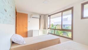 A hotel room with two beds, a window with a view of greenery, a desk, and an air conditioning unit.