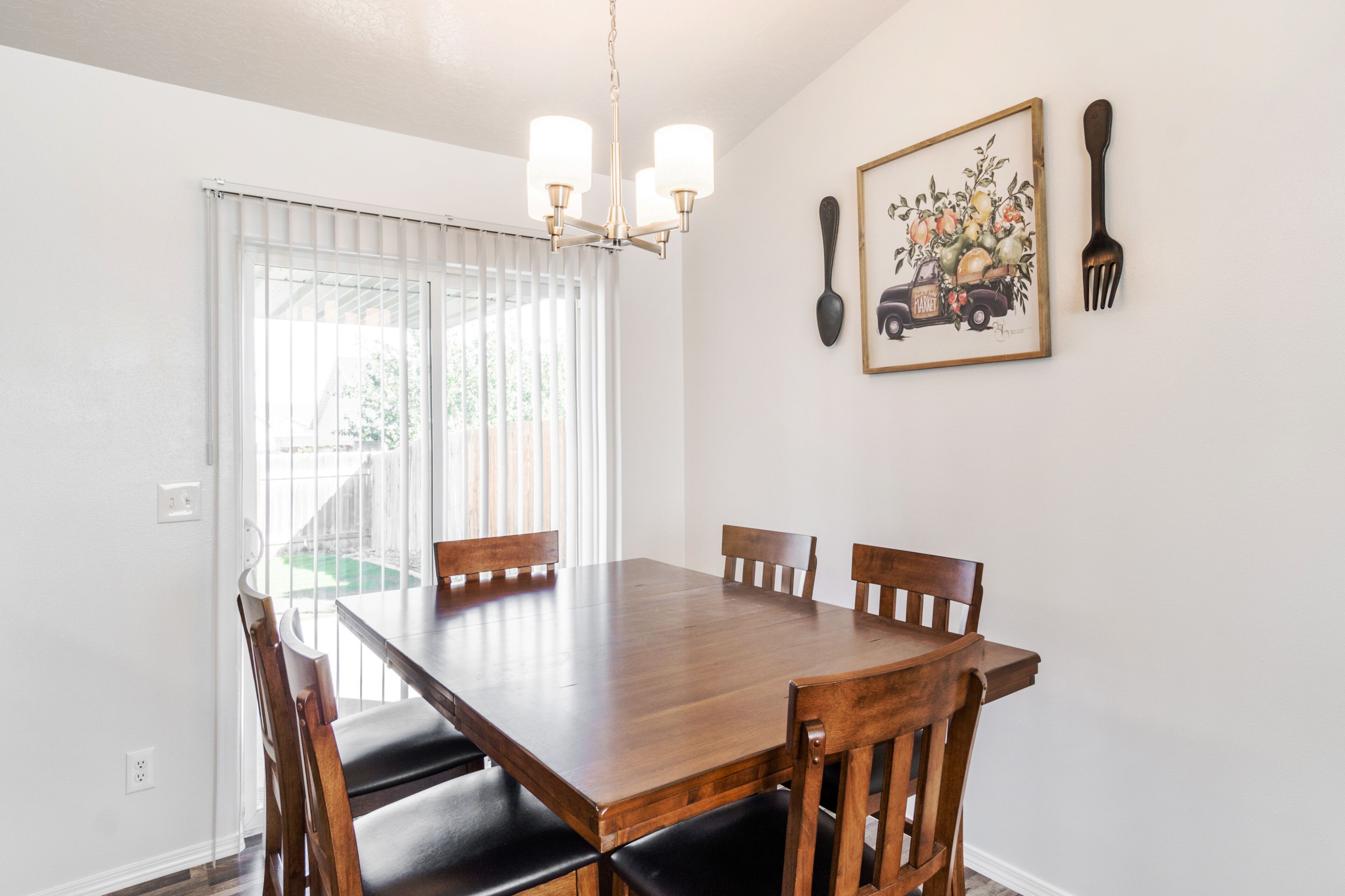 A dining area with a wooden table and chairs, a chandelier, a window with blinds, and a framed picture on the wall.