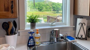 Uma cozinha com pia de aço inoxidável, janela com vista para uma paisagem verde, planta em vaso no parapeito e diversos utensílios e objetos de cozinha na bancada.