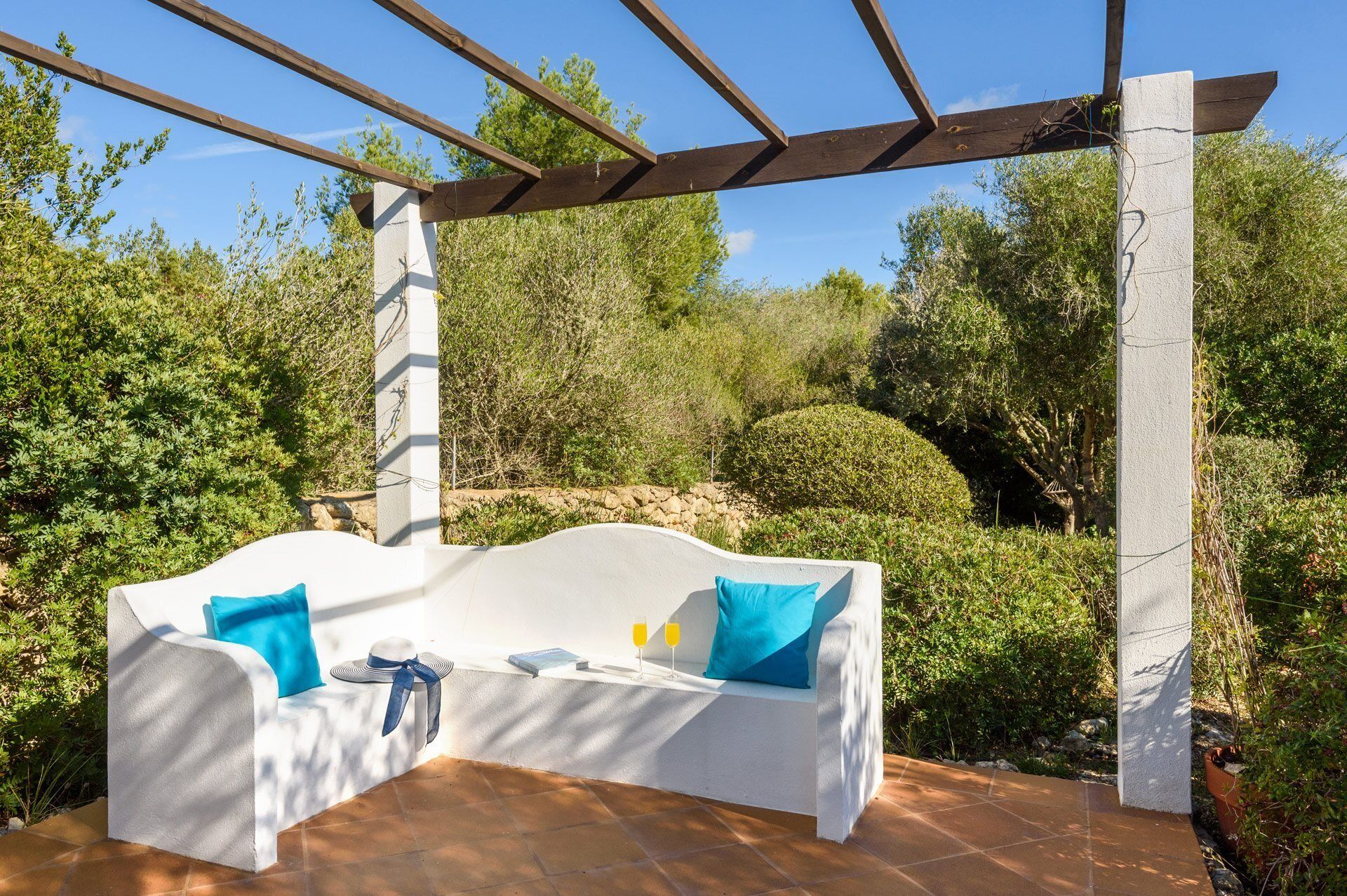 Un espace extérieur aménagé avec des sièges blancs, surmonté d’une pergola et entouré de verdure.