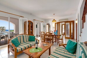 Un vaste espace de vie comprenant un ensemble de canapés, une table à manger et des chaises. Une porte ouverte offre une vue sur l’océan.