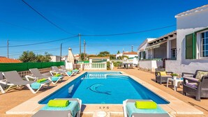 Ein Swimmingpool mit Liegestühlen und einem Tisch, umgeben von Gebäuden und blauem Himmel.