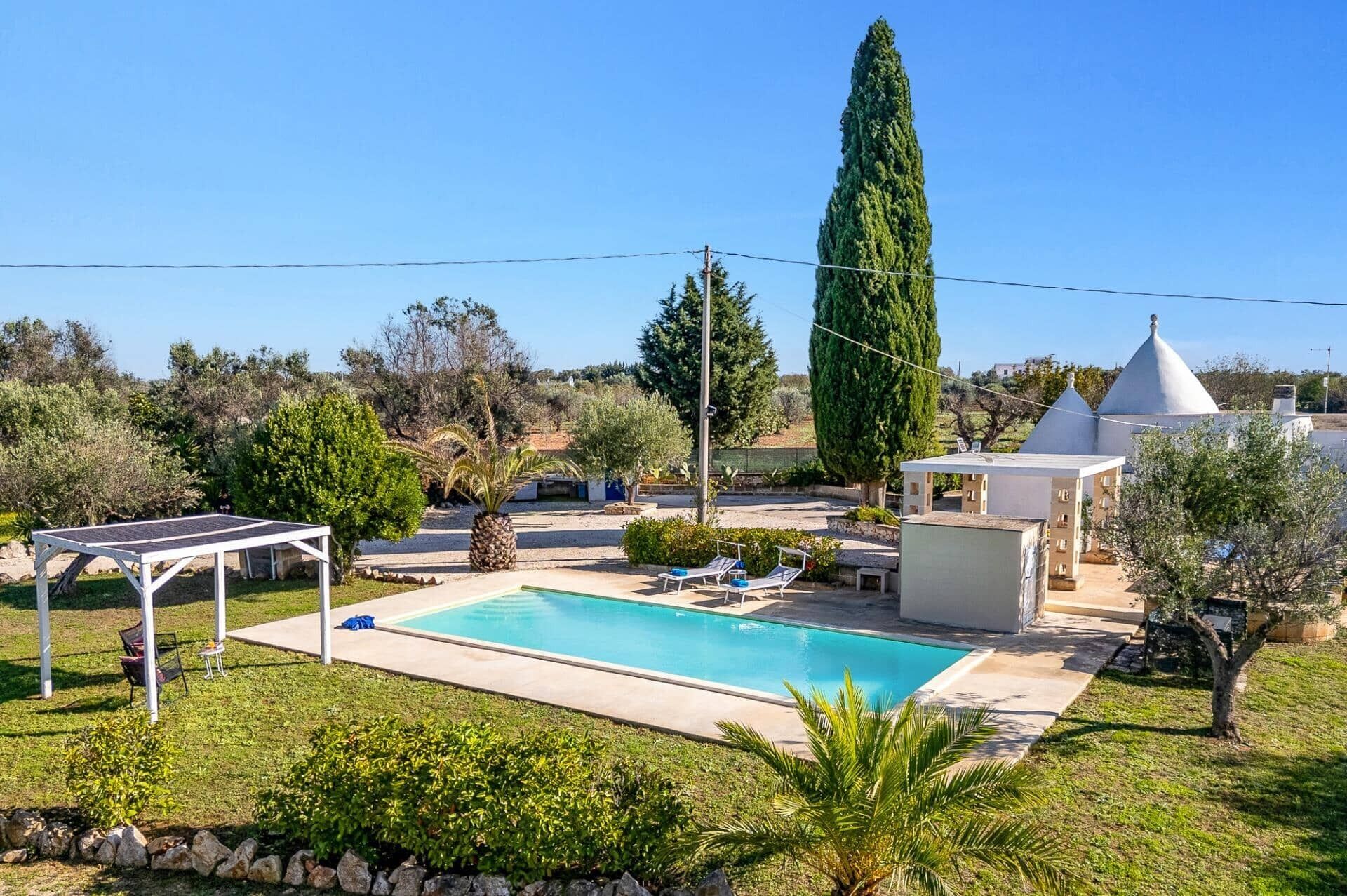 Ein Pool mit Liegestühlen, einer Pergola und einem hohen Baum in einem angelegten Garten.