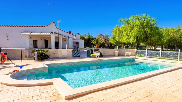Une piscine avec une terrasse carrelée, un patio couvert et un bâtiment blanc surmonté d’un toit recouvert de tuiles rouges.