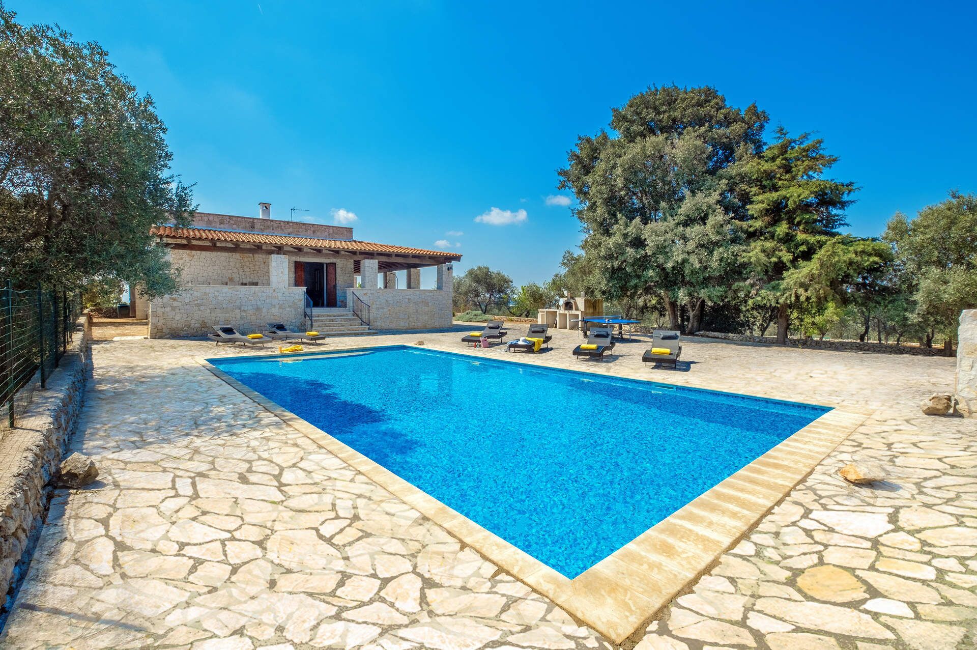 A rectangular swimming pool with stone tiles, surrounded by lounge chairs and a stone building with a tiled roof.