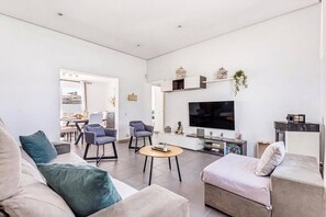 A modern living room with a flat-screen TV, sofas, a coffee table, and a dining area visible through an open door. - Villa Quimera by Villa Plus (Cala en Porter)