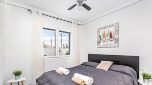 Un dormitorio con una cama grande, un ventilador de techo, una ventana con cortinas, un cuadro en la pared y dos mesitas con plantas.