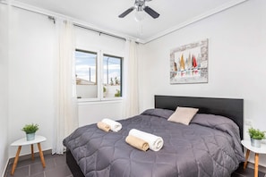A bedroom with a large bed, a ceiling fan, a window with curtains, a painting on the wall, and two small tables with plants.