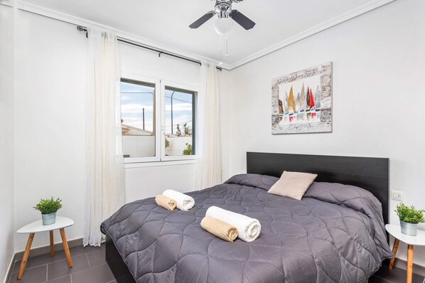 A bedroom with a large bed, a ceiling fan, a window with curtains, a painting on the wall, and two small tables with plants.