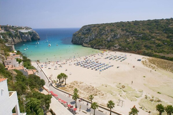 A beach with rows of sunbeds and umbrellas, a clear blue sea, and rocky cliffs in the background.
