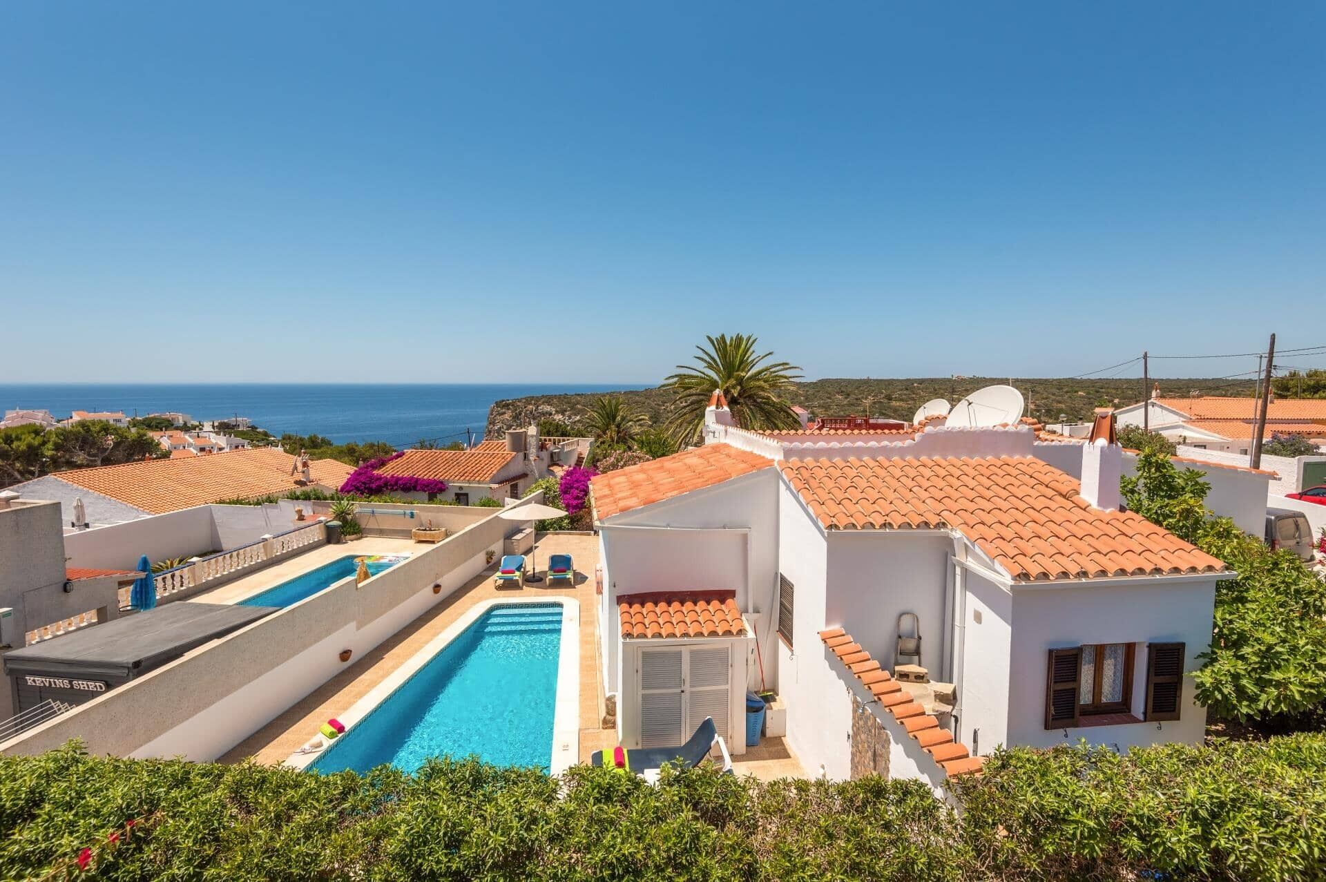 A residential area with a swimming pool, white buildings with red-tiled roofs, and a clear view of the sea.