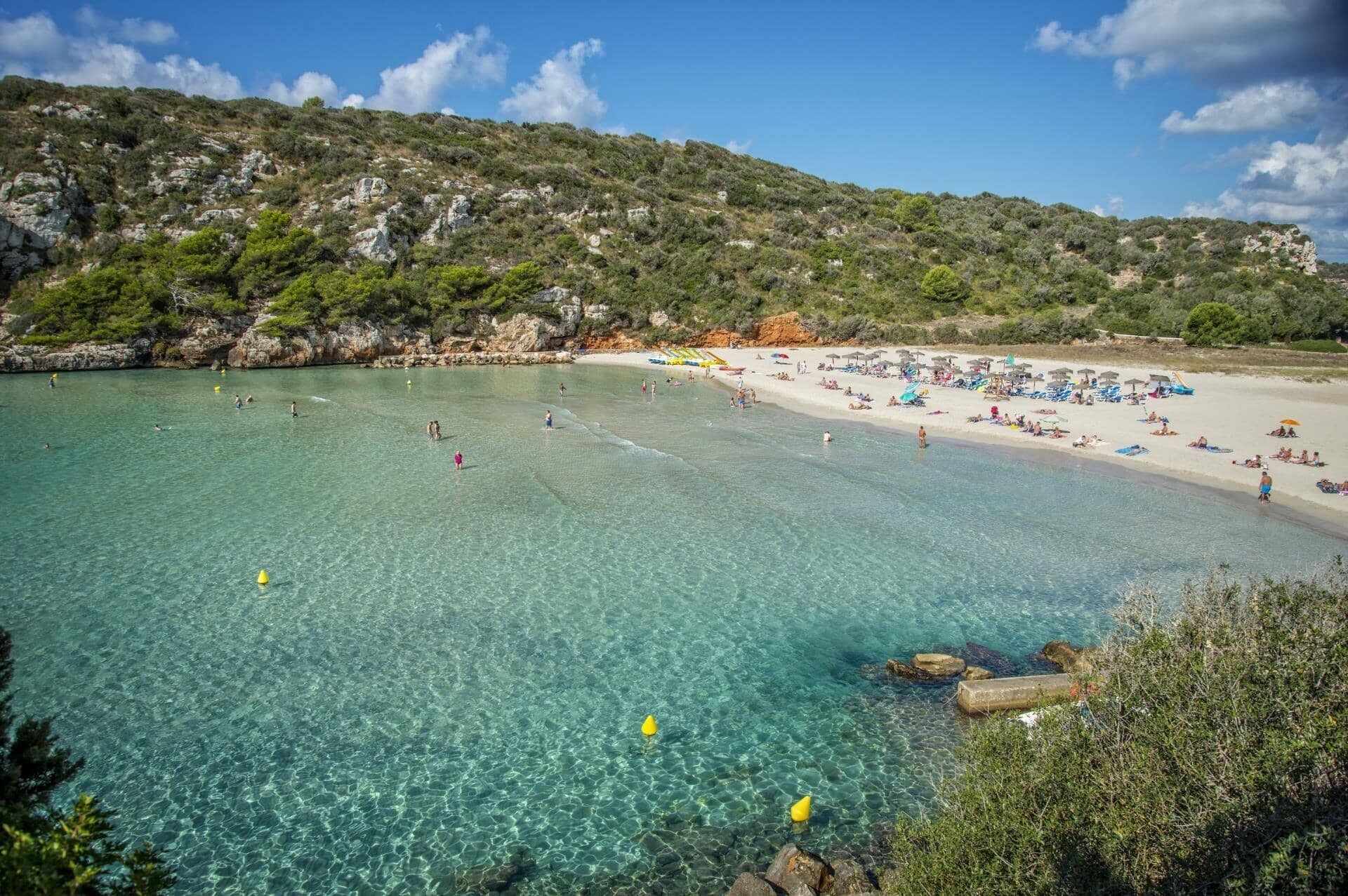 Een strand met helder turquoise water, rotsachtig terrein en een zandstrand met parasols en mensen.
