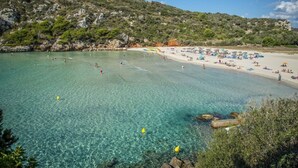Ein Strand mit klarem türkisblauem Wasser, felsigem Gelände und einem Sandstrand mit Sonnenschirmen und Badegästen.