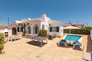 A white villa with a tiled patio, outdoor seating, a ping pong table, and a swimming pool.
