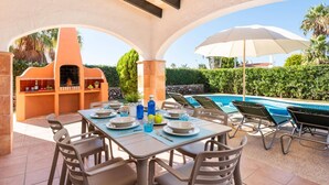 Un espace extérieur aménagé pour les repas, avec une piscine et des parasols, comprenant un barbecue et une table pour quatre personnes.