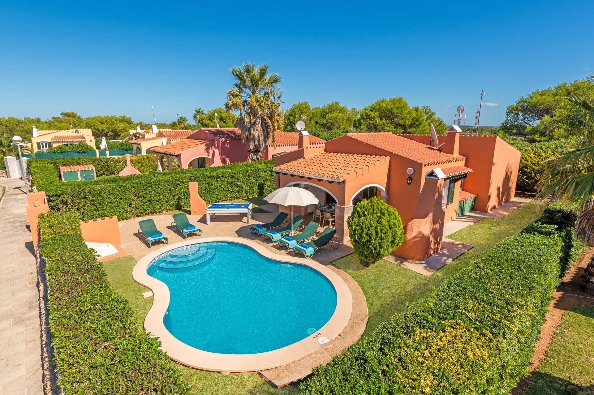 A pool area with lounge chairs and a sun umbrella, surrounded by well-maintained hedges and palm trees.
